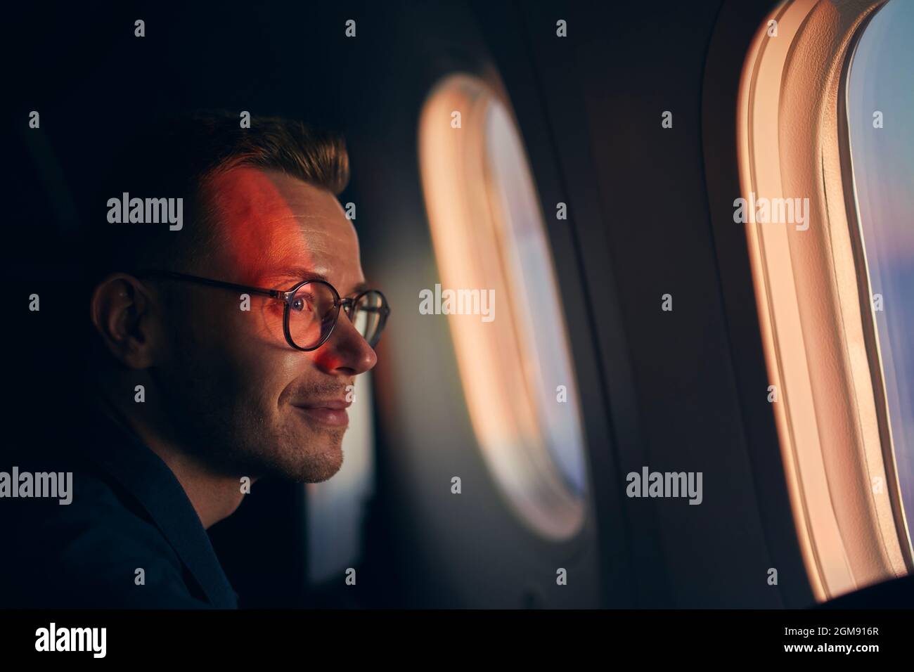 Junger Mann mit Brille, der mit dem Flugzeug reist. Passagier schaut während des Fluges bei Sonnenuntergang durch das Fenster. Stockfoto