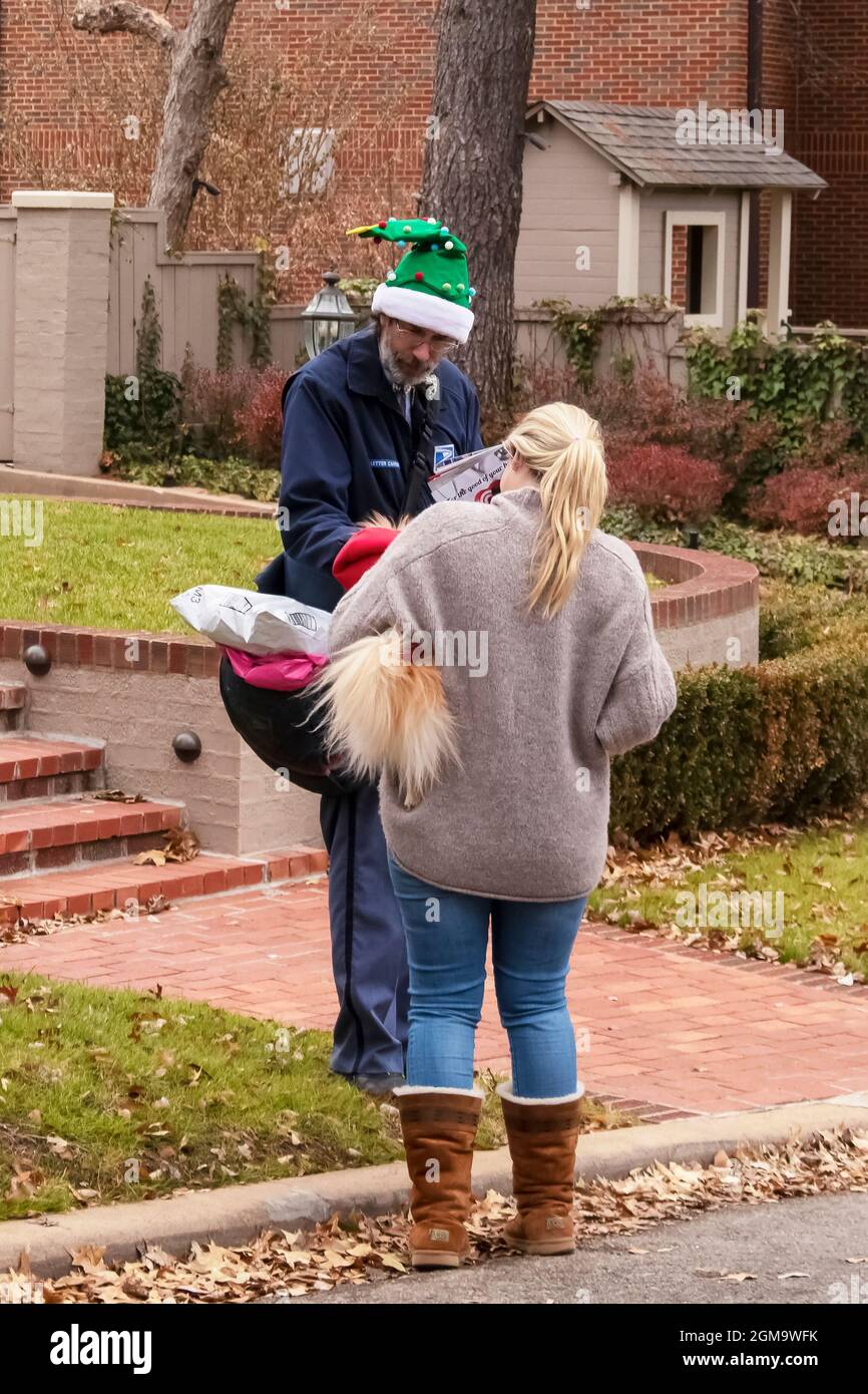 12-7-2018 Tulsa USA - Postmann trägt grünen Weihnachtselfenhut in seinen Runden zu PET Hund von ziemlich blonden Nachbarschaft Frau in Pullover und gehalten pausiert Stockfoto