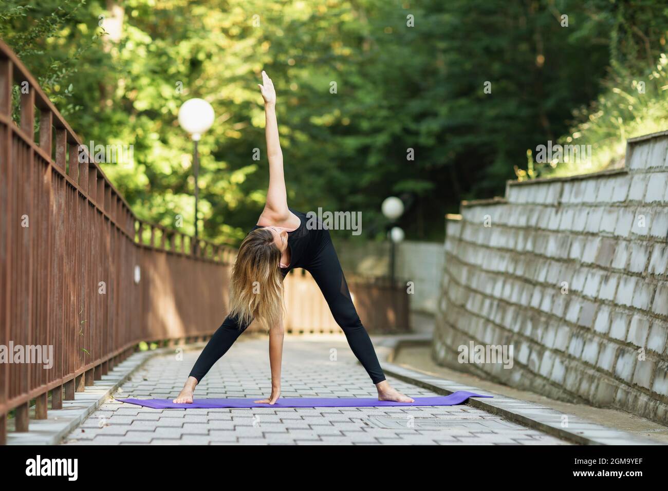 Eine Frau, die Yoga praktiziert, eine Drehübung Parivritta Padottanasana macht, an einem Sommermorgen im Park trainiert, auf einer Matte stehend Stockfoto