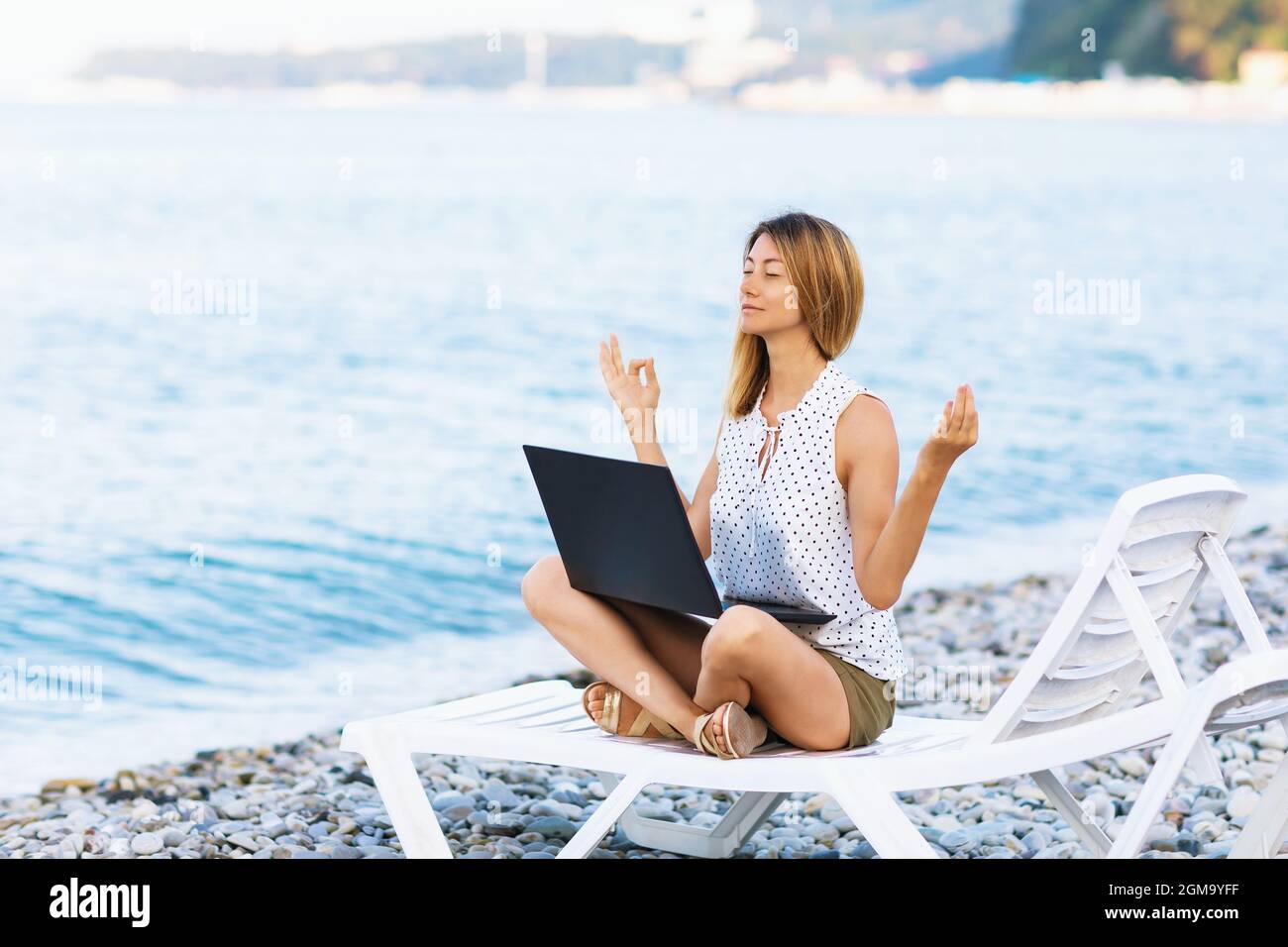 Eine ruhige Frau sitzt auf einer Sonnenliege nahe dem Meer, wird von der Arbeit am Computer abgelenkt, meditiert und entfernt negative Emotionen, kombiniert Remote Stockfoto
