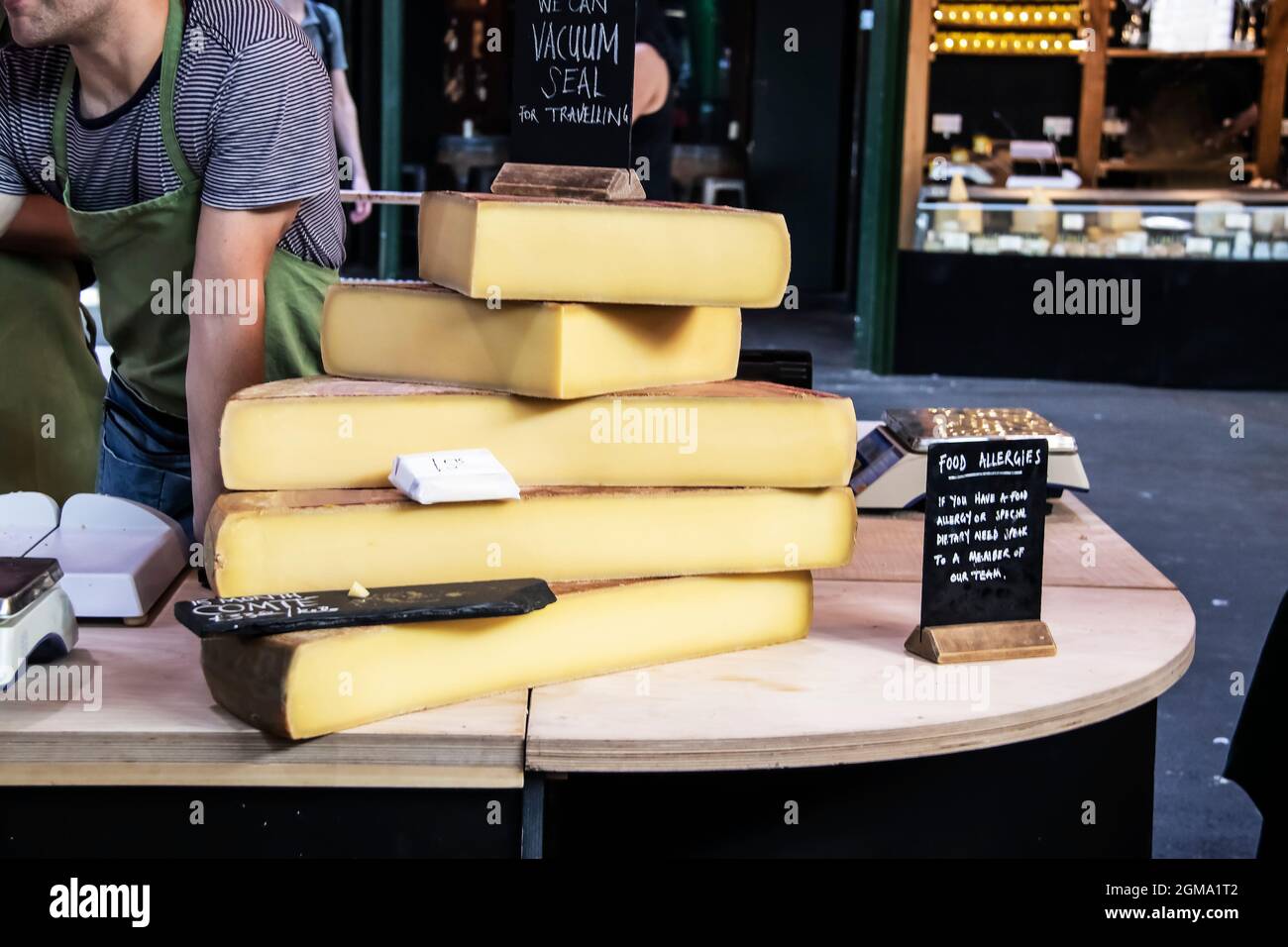 Große Käseplatten auf einem Holztisch auf einem Markt, auf dem sich der Arbeiter über das Gespräch mit dem Kunden lehnte, und auf Tischen mit Waage und Salbe Schilder für Lebensmittelallergien Stockfoto