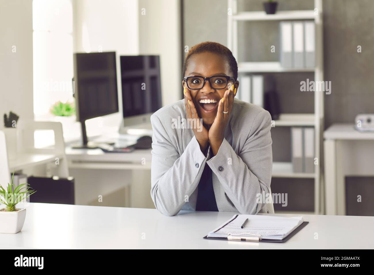 Aufgeregte schwarze Frau, die am Schreibtisch sitzt und mit fröhlicher Gesichtsausdruck auf die Kamera schaut Stockfoto