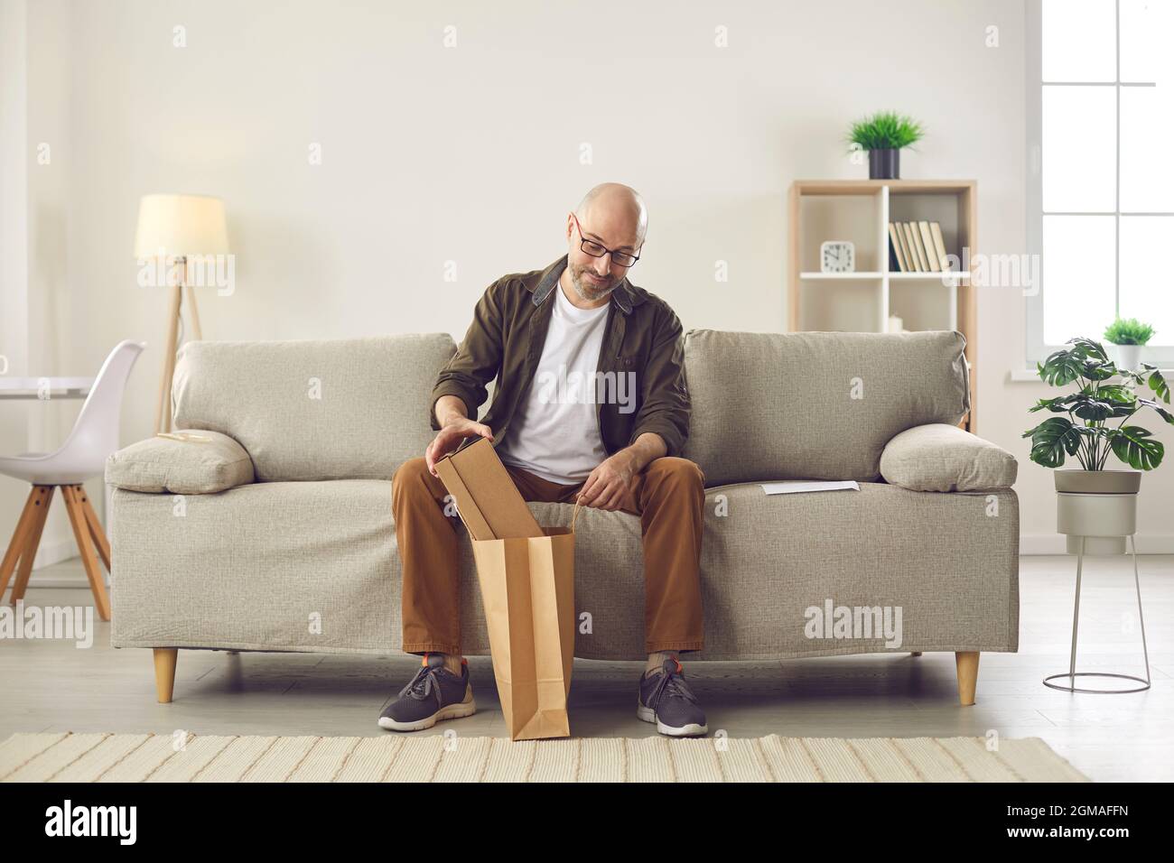 Ein glücklicher Mann, der zu Hause auf dem Sofa sitzt, nimmt den Karton aus dem Papierlieferbeutel Stockfoto