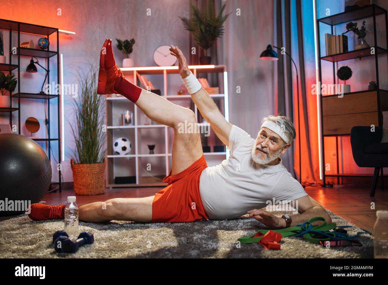Aktiver älterer Mann in Sportkleidung, die auf dem Boden liegt und Beine hebt. Kaukasischer bärtiger Mann, der abends Fitnessübungen macht. Hausarbeit und Rentenkonzept. Stockfoto