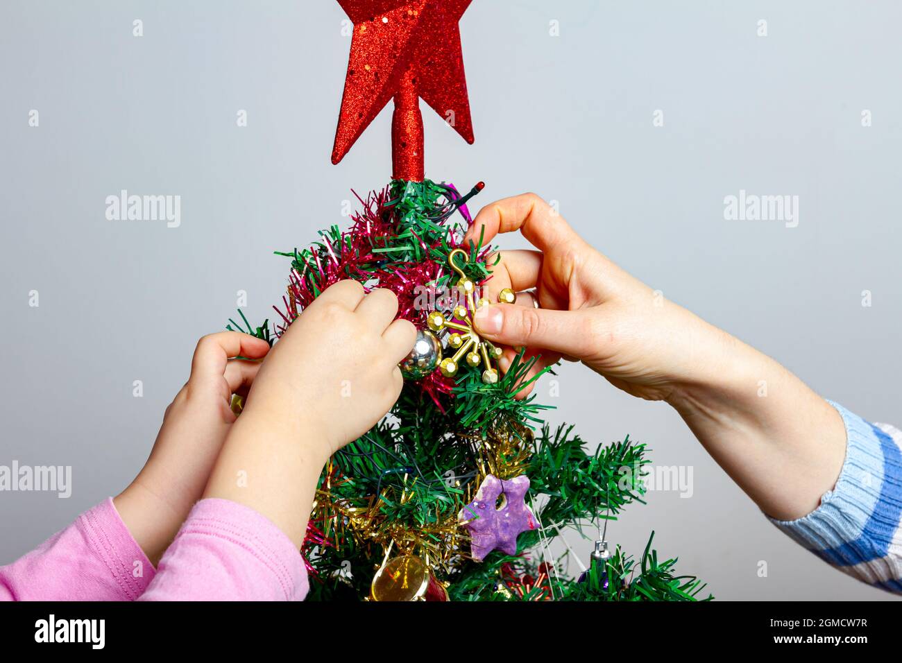 Nahaufnahme der Hände als Mutter ihrer Tochter eine Hand gibt, um glänzende Dekoration mit Quaste auf Kunststoff Evergreen, Kiefer, Baum für Heiligabend zu platzieren. Stockfoto