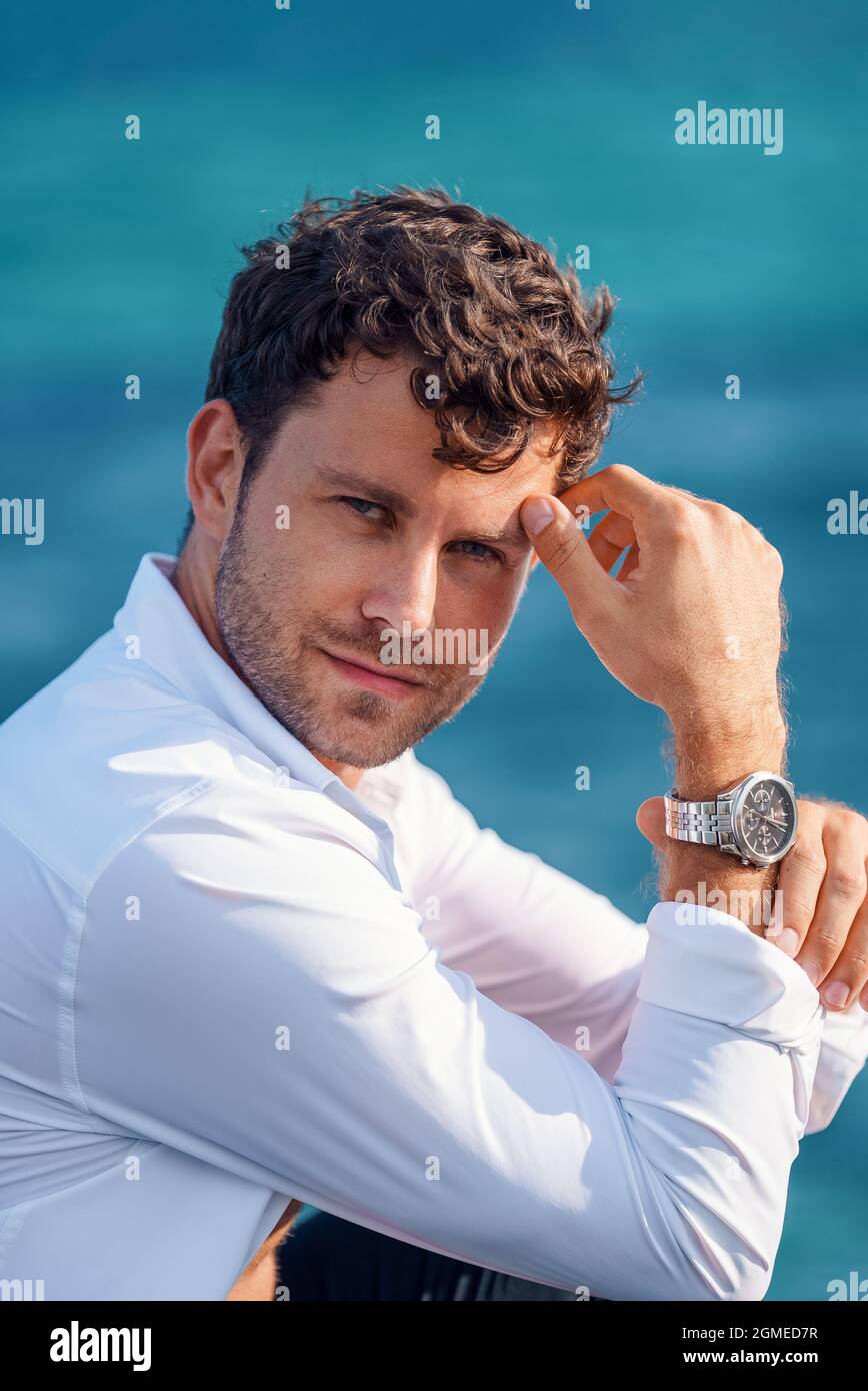 Seitenansicht eines selbstbewussten, hübschen Mannes mit weißem Hemd und Armbanduhr, der sich an der Hand lehnt und die Kamera am sonnigen Tag vor verschwommenem Meereshintergrund anschaut Stockfoto