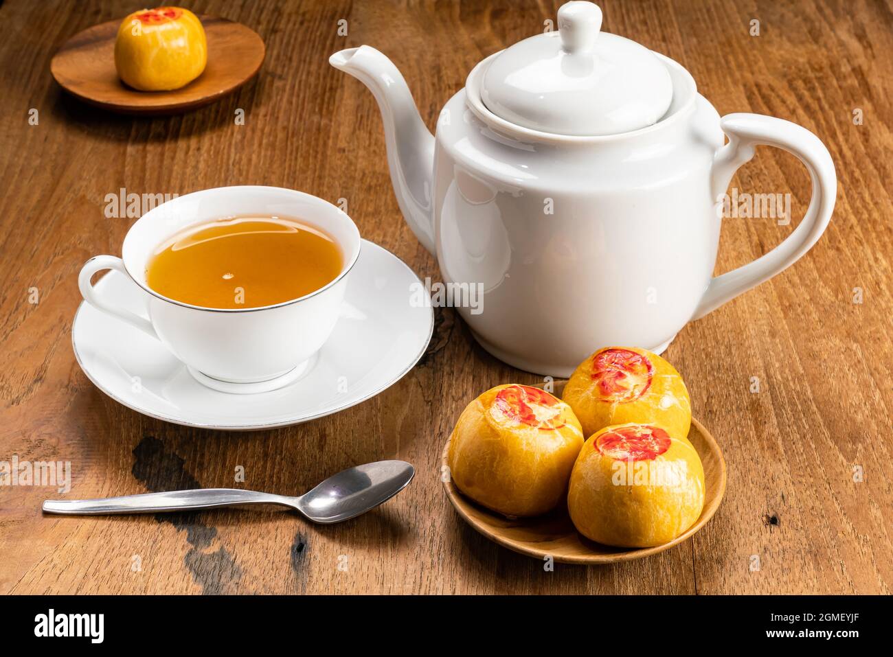 Blick auf süßes chinesisches Gebäck oder Mondkuchen, gefüllt mit süßer Mungbohnenpaste und einer weißen Tasse heißen Tees aus Keramik mit Wasserkocher und Metalllöffel auf Holz-T Stockfoto