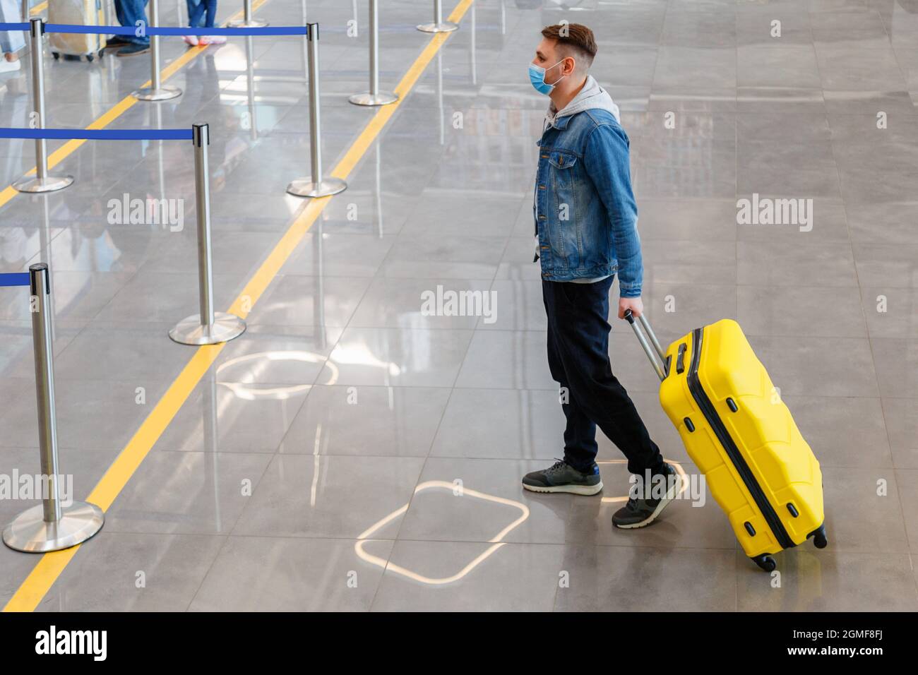 Mann mit medizinischer Gesichtsmaske vor dem Check-in-Schalter im Flughafeninneren während der Zwangsabsperrung. Urbane Freizeitreisende mit Kofferraumbeförderung Stockfoto