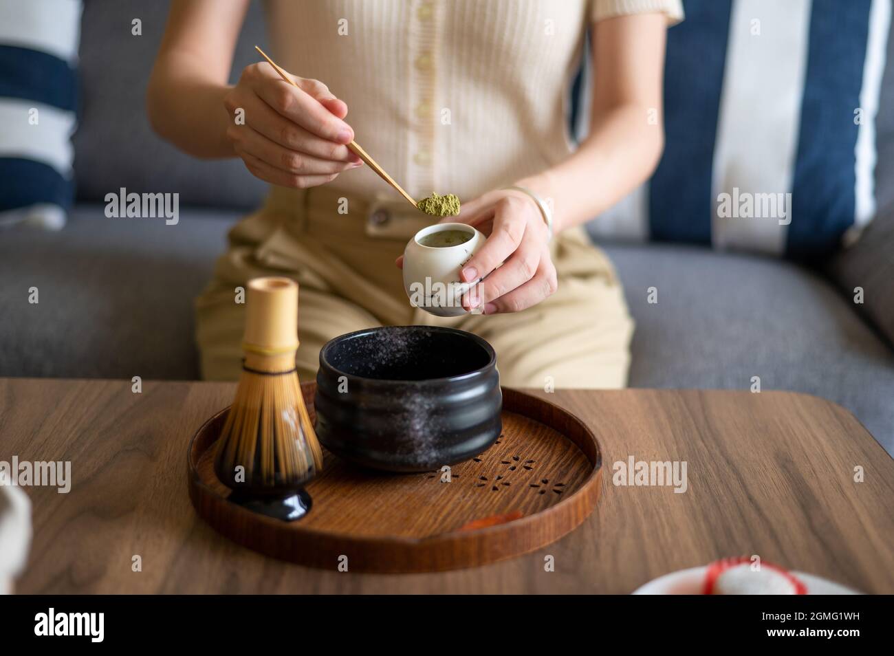 Frau, die zu Hause einen japanischen Matcha-Grüntee mit traditionellem Bambus-Set für Teezeremonien zubereitet, während sie zu Hause auf dem Sofa sitzt. Stockfoto