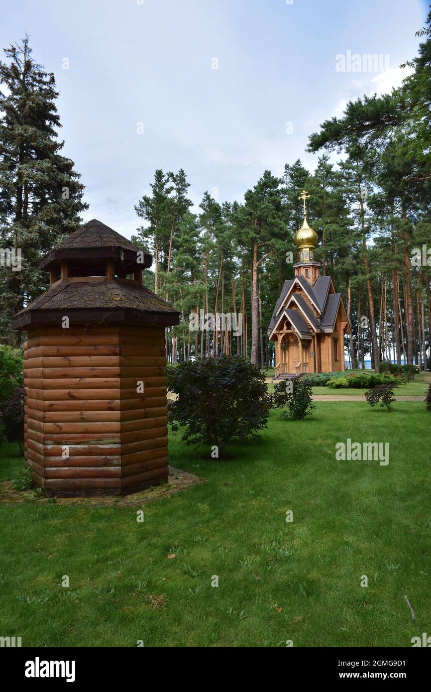 In einem Kiefernsommerwaldpark gibt es eine kleine hölzerne christliche Kirchenkapelle. Im Vordergrund ist ein Holzhaus ohne Fenster. Stockfoto