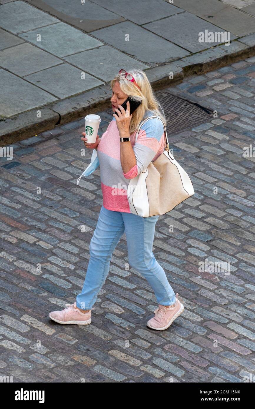 Frau, die eine gepflasterte Hauptstraße entlang geht und mit einem Mobiltelefon spricht, eine Tasse Starbucks Kaffee mit einer Gesichtsmaske am Handgelenk hält, 2021 UK Stockfoto