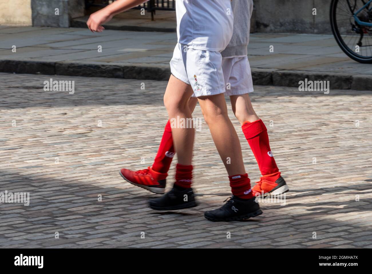 Zwei Schuljungen, die in Sportkleidung eine gepflasterte Straße entlang gehen - Nahaufnahme ihrer Beine in roten Fußballsocken und weißen Shorts Stockfoto