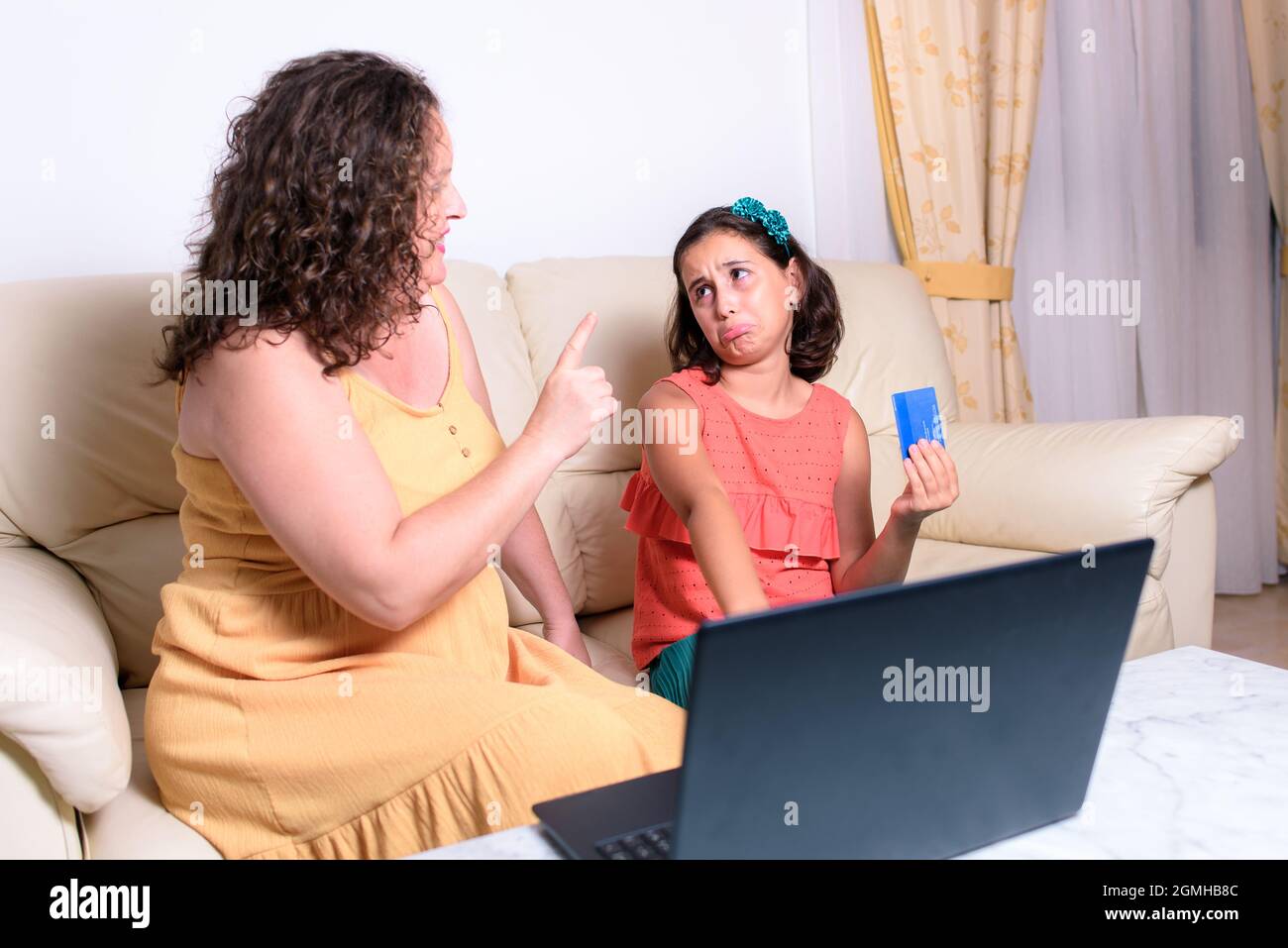 Das brünette Mädchen sitzt zu Hause auf der Couch und macht ein klägliches Gesicht, um online etwas für sie zu kaufen, aber ihre Mutter zeigt mit dem Finger auf sie und sagt nein Stockfoto
