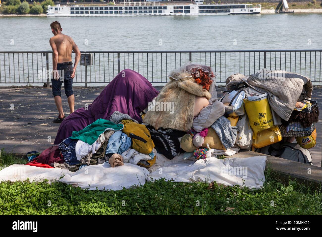 Obdachlose mit ihren Besitztümern am Rheinufer, Köln, Deutschland. Ein Mann ohne Hemd, eine Frau hockt mit einem Pelzumhang auf ihrer Tasche Stockfoto