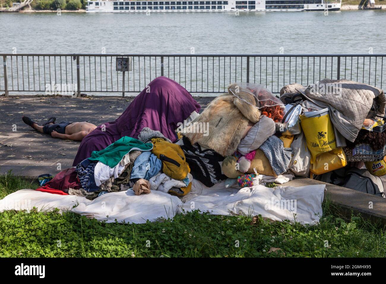 Obdachlose mit ihren Besitztümern am Rheinufer, Köln, Deutschland. Ein Mann liegt auf dem Bürgersteig, eine Frau hockt mit einem Pelzumhang auf ihm Stockfoto