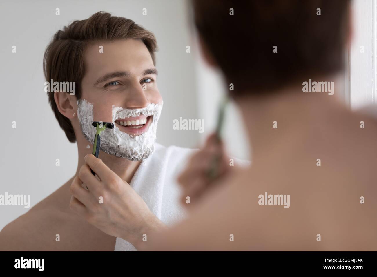 Hübscher junger Mann, der sich sein Gesicht rasiert und in einen Spiegel schaut Stockfoto