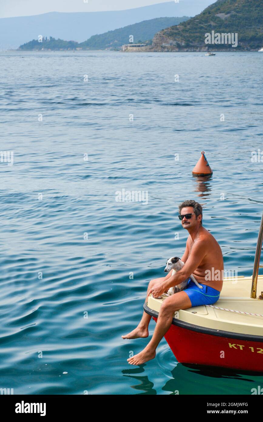 Ein Mann mit seinem kleinen Hund sitzt am Bug eines kleinen Motorbootes auf dem ruhigen Wasser der Kotor Bay. Stockfoto