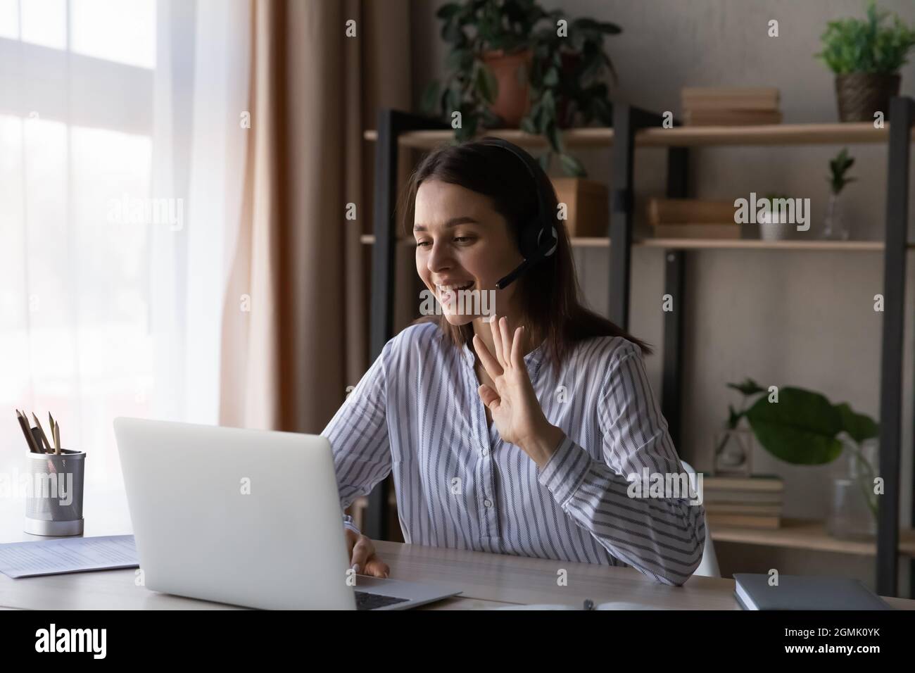 Lächelnde Frau im Headset winkend mit der Hand, mit dem Laptop, online chatend Stockfoto