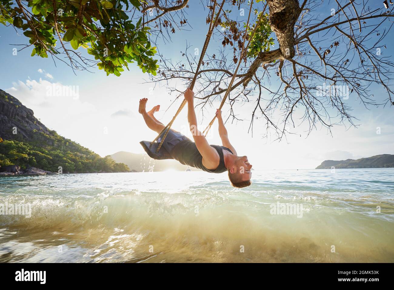 Glücklicher Mann auf der Schaukel, der den Urlaub genießt. Wunderschöner Sonnenuntergang am Strand auf den Seychellen. Stockfoto