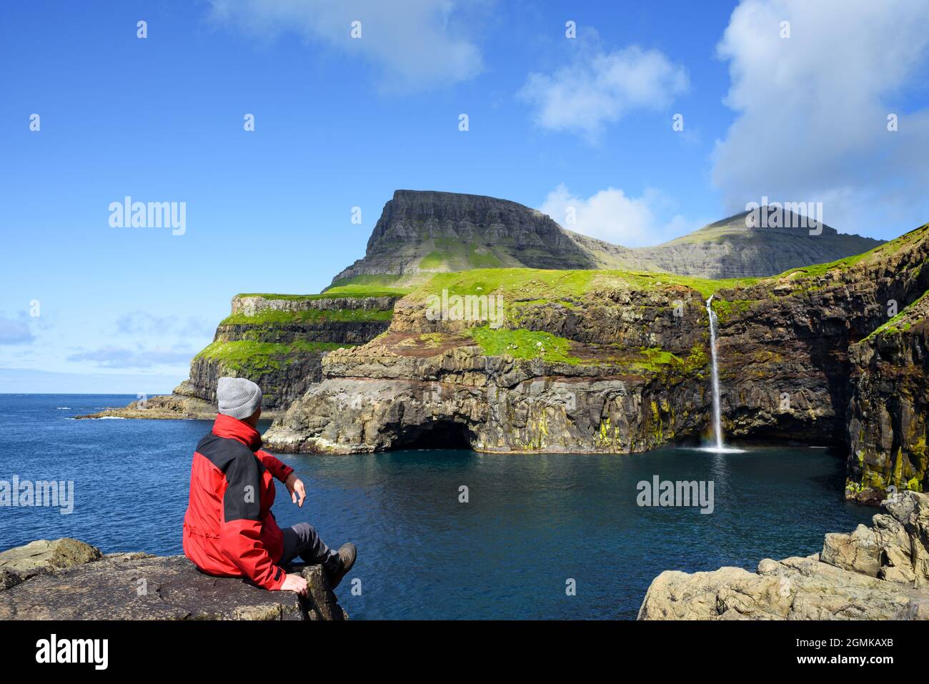 Ein Tourist in einer roten Jacke blickt auf einen Mulafossur-Wasserfall im Dorf Gasadalur auf den Färöer Inseln Stockfoto