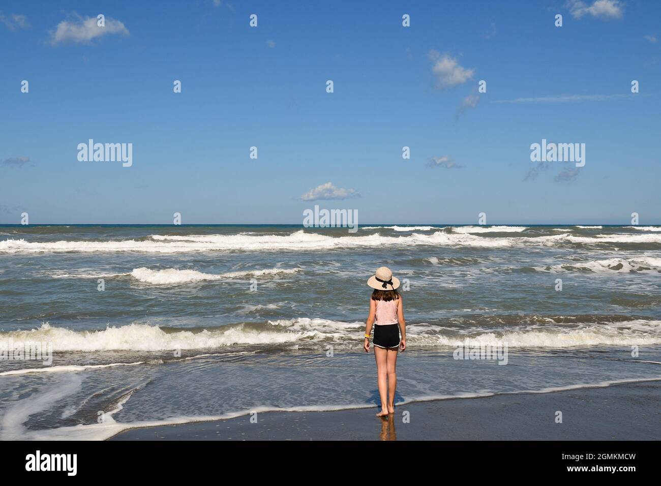 Rückansicht eines kleinen Mädchens (10 Jahre alt) mit Strohhut, das im Sommer am Ufer des rauhen Meeres steht, Marina di Castagneto Carducci Stockfoto