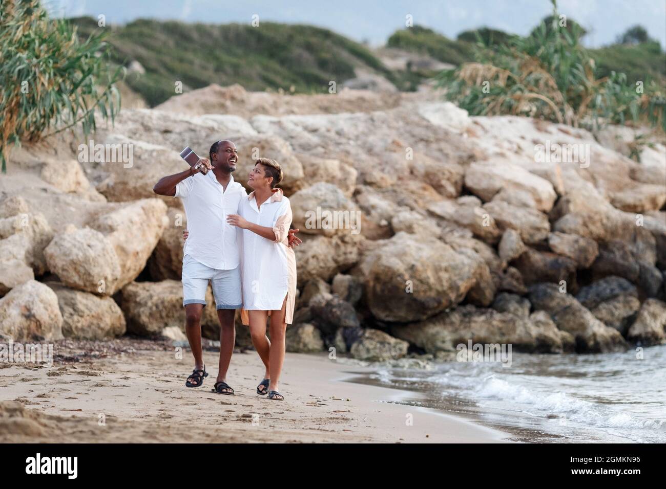 Fröhliches gemischtes Rennen, Paar mittleren Alters, das sich umarmt, während man am Strand mit einer Gitarre läuft. Mann mit Gitarre und Frau. Stockfoto