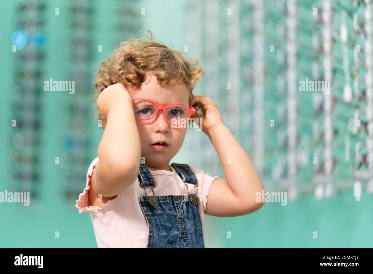 Ein blondes kleines Mädchen, das in einem Optikerladen eine Brille anprobiert. Stockfoto