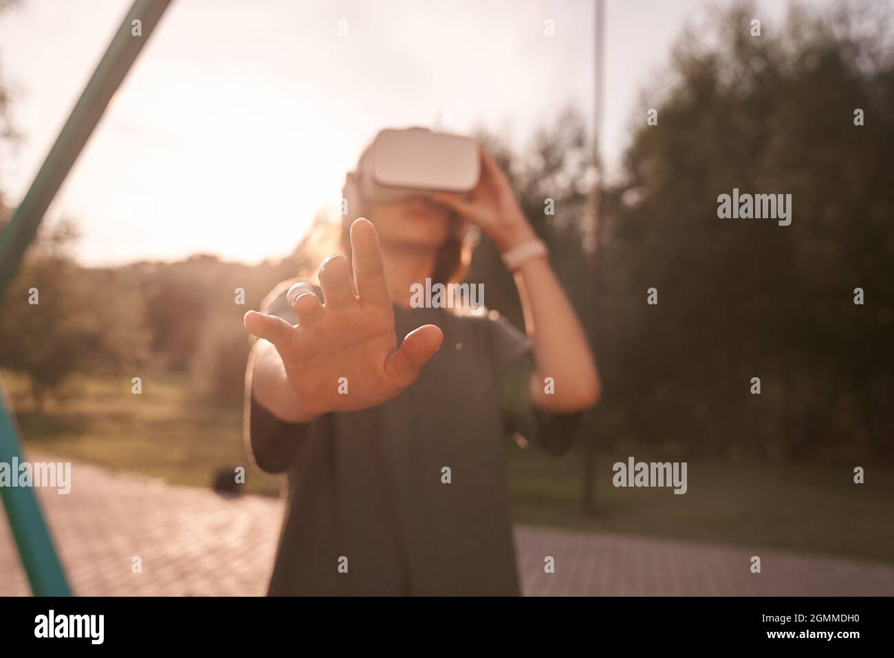 Nahaufnahme eines Mädchens, das mit den Händen eine Virtual-Reality-Brille auf dem Kopf hält und auf einem Hintergrund von grünen Bäumen steht. Er reicht eine Hand an die Kamera. Hochwertige Fotos Stockfoto