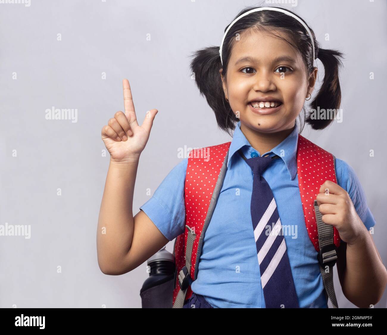 Porträt eines glücklich lächelnden indischen Mädchen Schüler in blauer Schuluniform mit roter Tasche zeigt auf leeren Bereich auf grauem Hintergrund Stockfoto