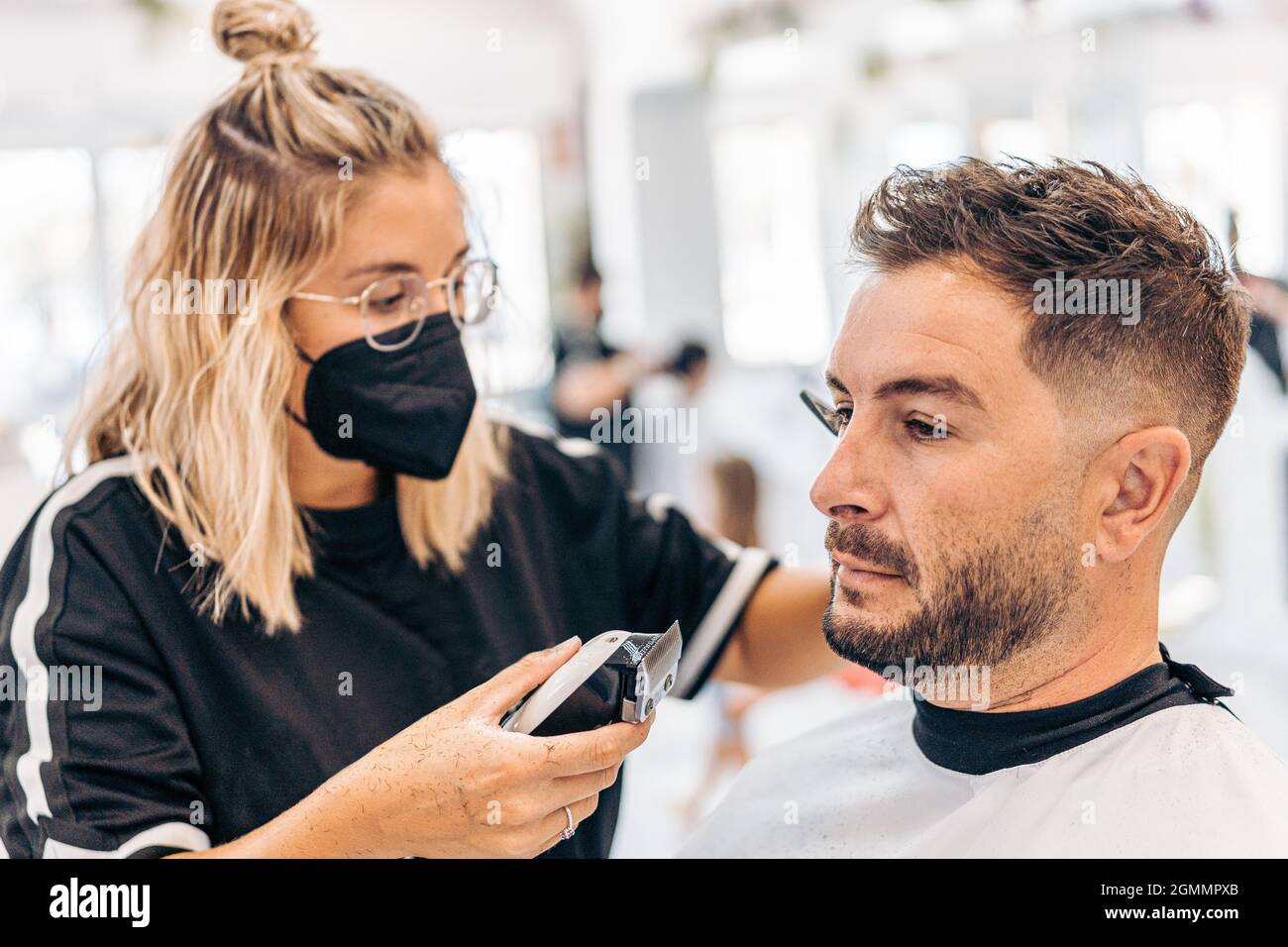 Frau mit Maske, die einen Mann mit einer elektrischen Maschine in einem Friseurladen rasiert Stockfoto