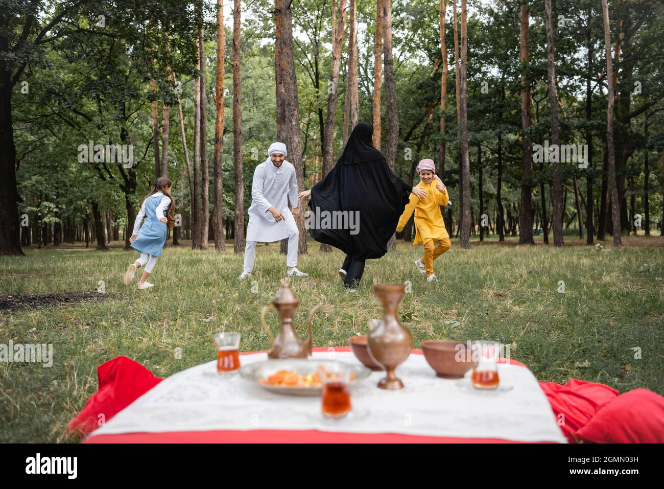 Positive arabische Familie, die während des Picknicks im Park mit Kindern in der Nähe von verschwommenem Essen spielt Stockfoto