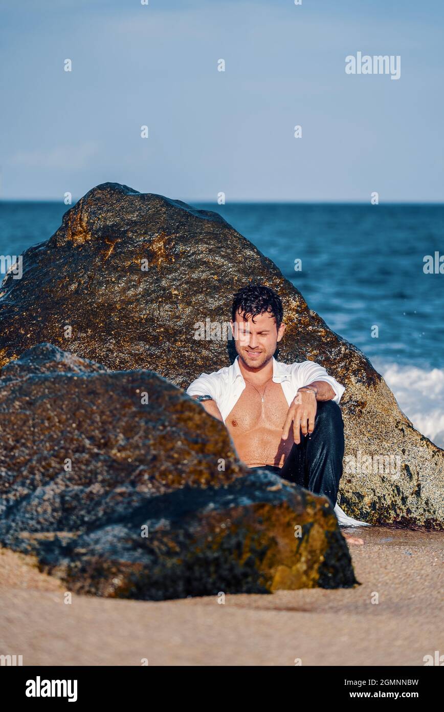 Inhalt schöner Mann mit feinem Körper und in weißem Hemd, der an sonnigen Tagen in der Nähe von nassem Felsen am Meer sitzt Stockfoto