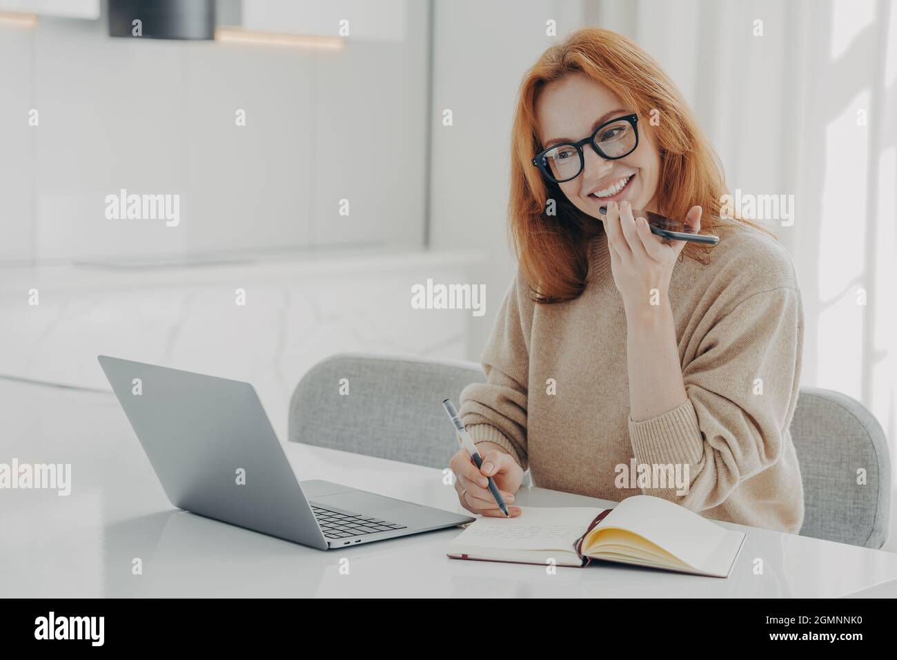 Positive Ingwer-Frau in legerer Kleidung nimmt während der Arbeit am Laptop Audio-Nachrichten oder Smartphones auf Stockfoto