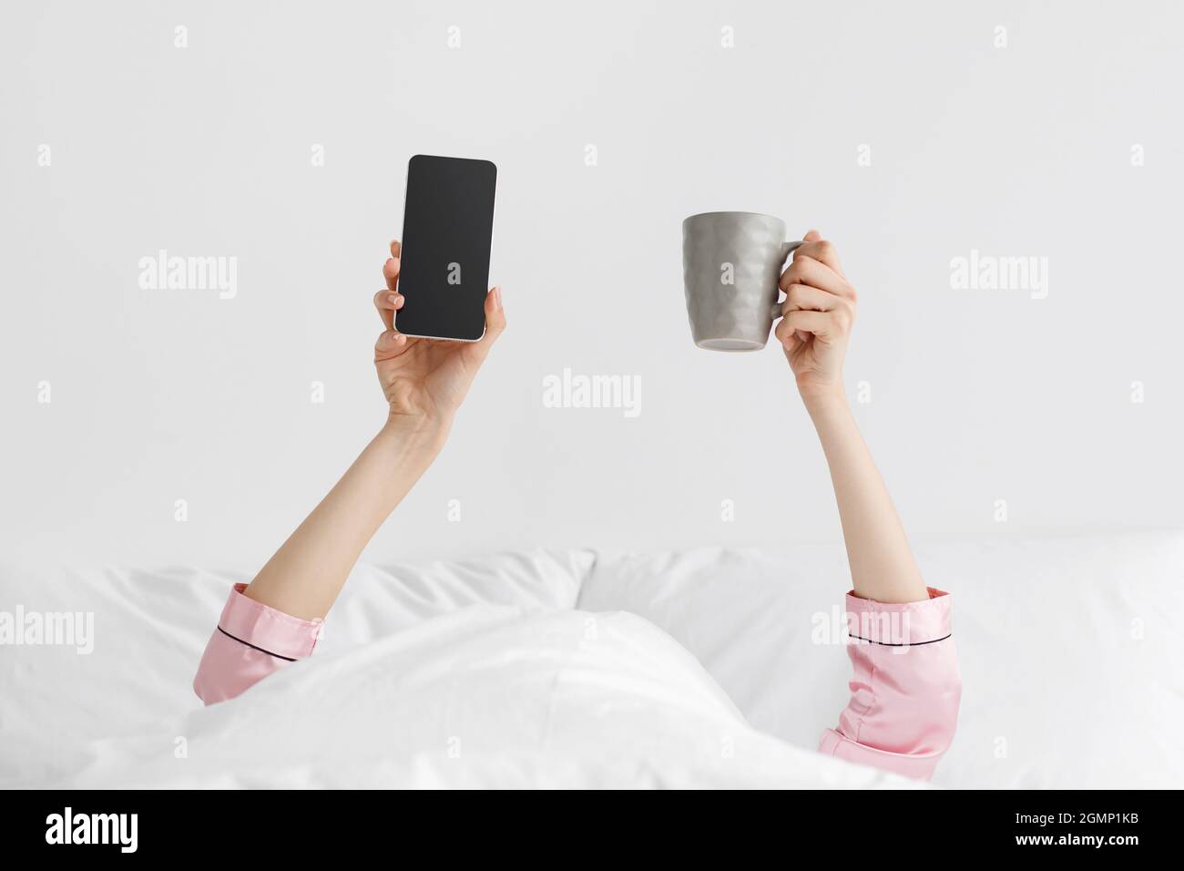 Hände einer tausendjährigen kaukasischen Dame in rosa Pyjamas ragen aus der weißen Decke und halten eine Tasse Kaffee und ein Smartphone Stockfoto