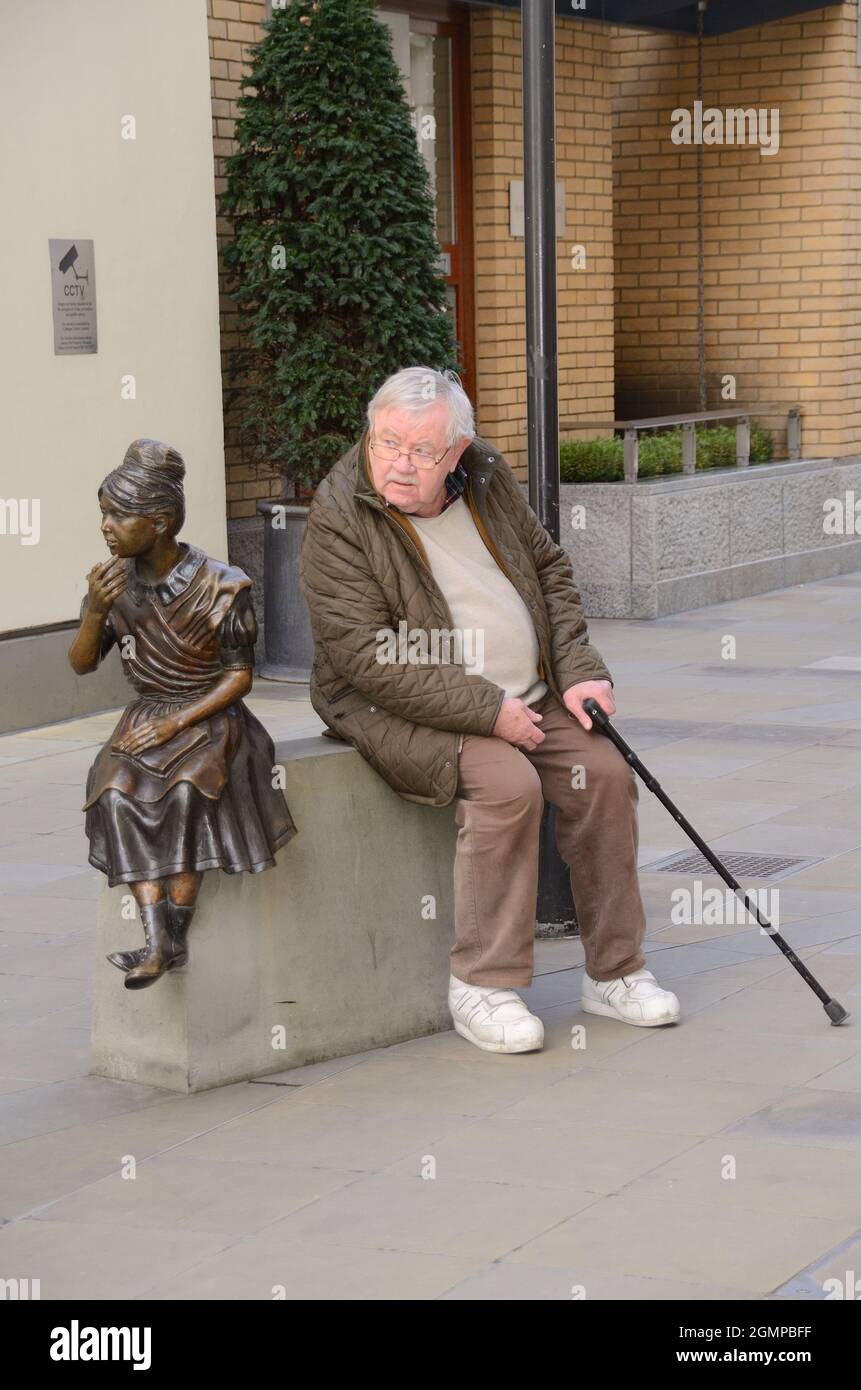 Der alte Mann saß neben der Statue des Mädchens auf einem Sockel. London, Großbritannien. Stockfoto