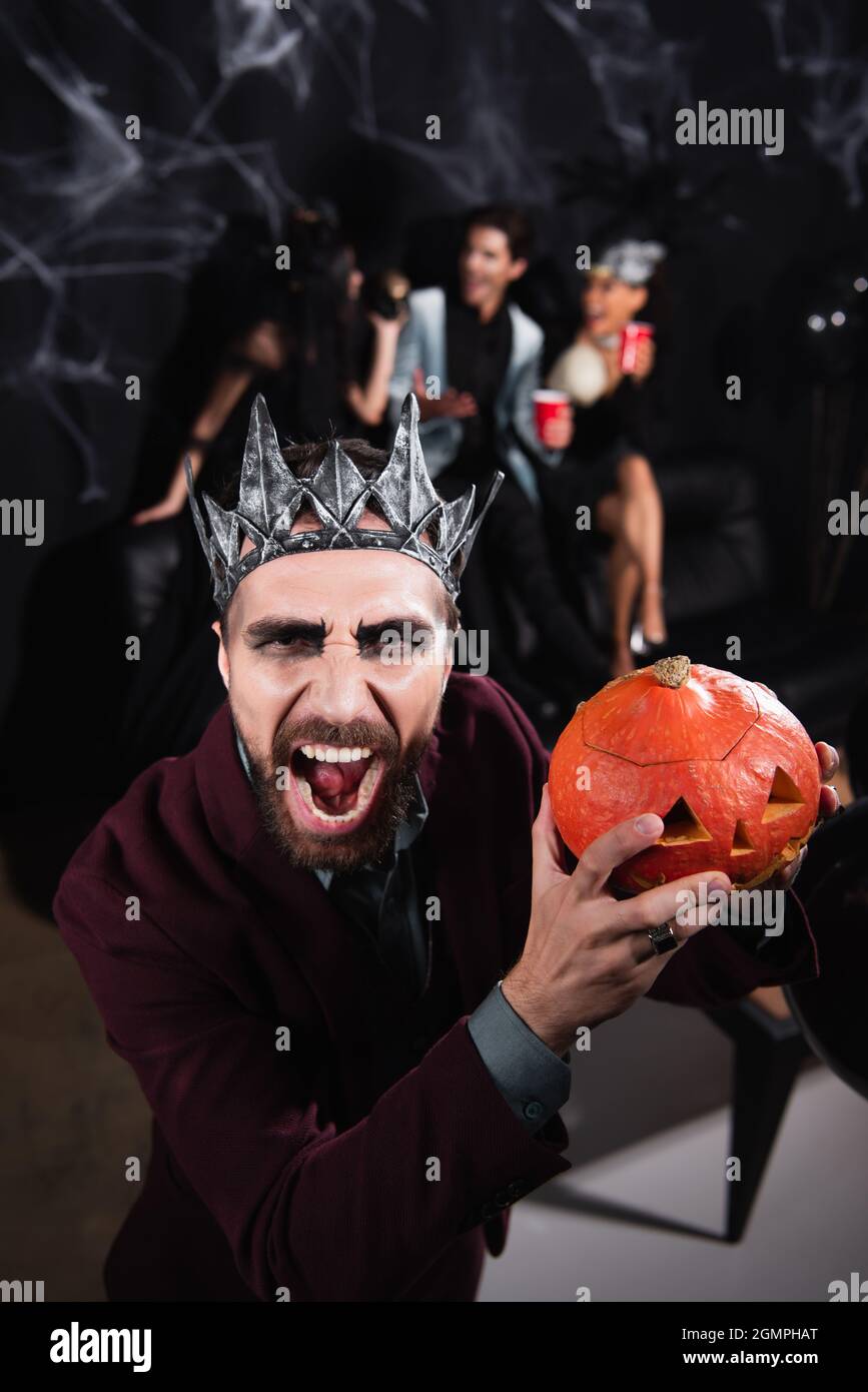 Wütender Mann in der Vampirkönigskrone knurrt vor der Kamera, während er den halloween-Kürbis auf Schwarz hält Stockfoto