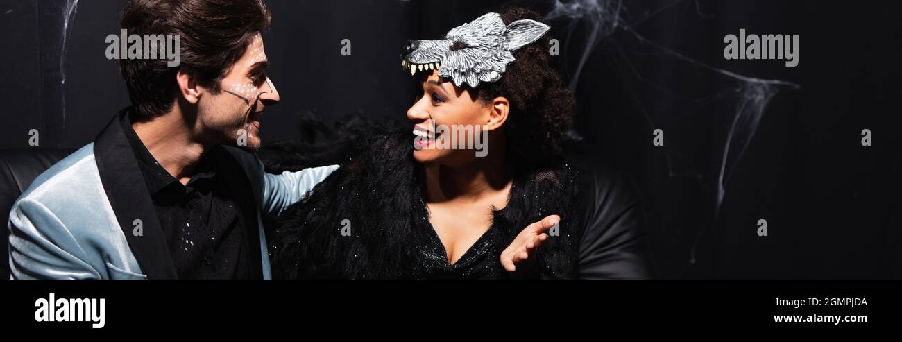 Glückliche afroamerikanische Frau in Wolf Maske im Gespräch mit Mann in halloween Make-up, Banner Stockfoto