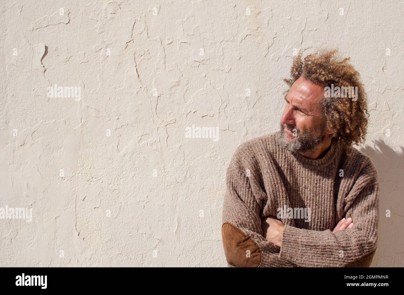 Der 50-60-jährige ältere Mann mit langen Haaren, die an einer weißen Wand gelehnt sind, blickt zur Seite, am Horizont mit gekreuzten Armen Stockfoto