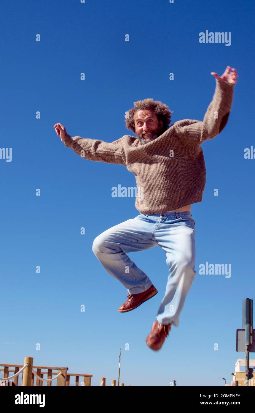 Lächelnder, 50-60 Jahre alter Mann mit lockigem Haar liegt in der Luft für einen Sprung und hebt seine Arme Stockfoto