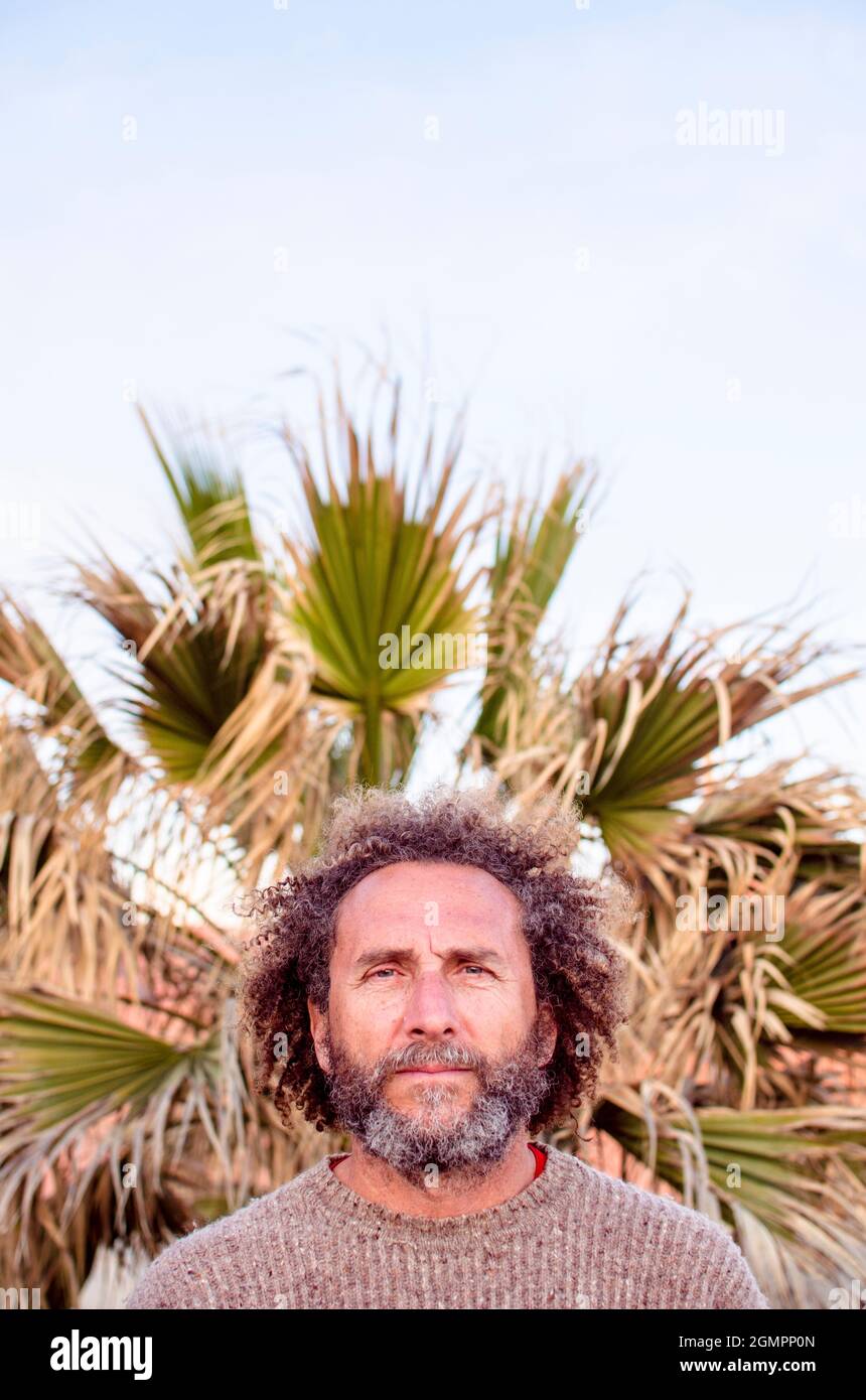 Mann mittleren Alters betrachtet die Kamera, seine krauses Haar fügt sich mit einer Palme im Hintergrund Stockfoto