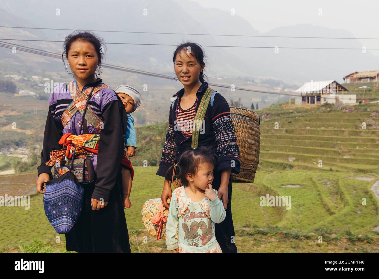 Sapa, Vietnam - 15. April 2016: Schwarze Hmong-Minderheitsfrau trägt Kind auf dem Rücken, während sie in den Bergen arbeitet und geht. Traditionelle Schlinge Stockfoto