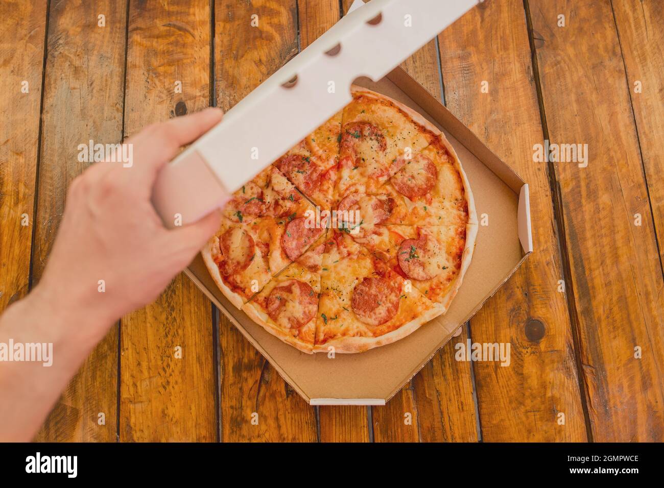 Die Hand des Typen öffnet eine Pappschachtel Pizza vor dem Hintergrund eines Holztisches. Köstliches Fast Food. Stockfoto