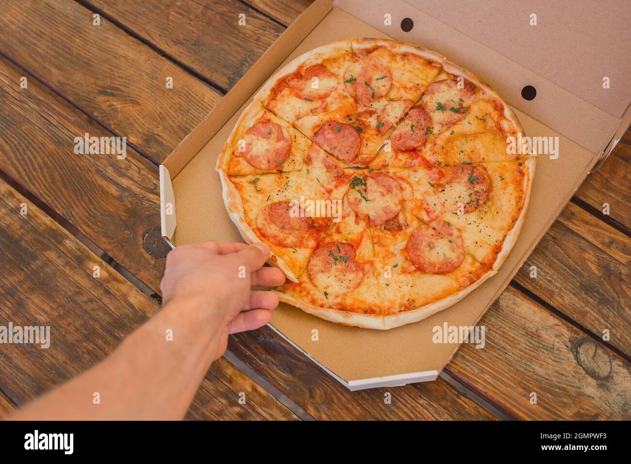 Die Hand des Mannes nimmt eine Pizza in einer Pappschachtel vor dem Hintergrund eines Holztisches. Köstliches Fast Food. Stockfoto