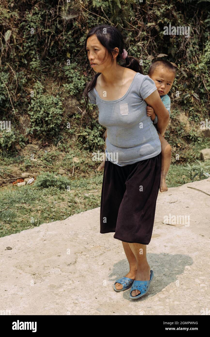 Sapa, Vietnam - 15. April 2016: Schwarze Hmong-Minderheitsfrau trägt Kind auf dem Rücken, während sie in den Bergen arbeitet und geht. Traditionelle Methode Stockfoto