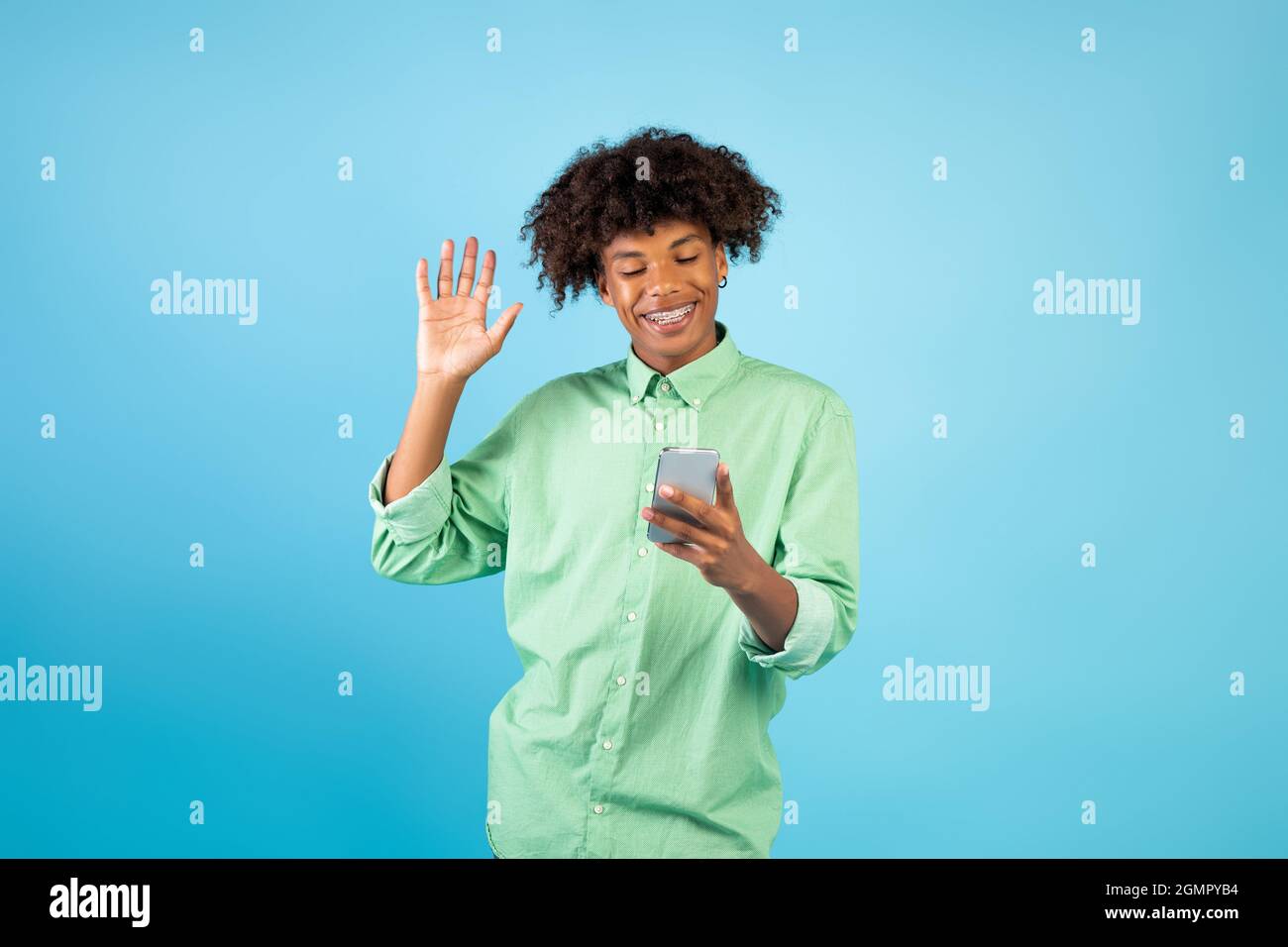 Glücklicher schwarzer Teenager-Kerl mit Smartphone macht Videoanruf, winkende Hand auf Handy-Kamera, blauer Hintergrund Stockfoto