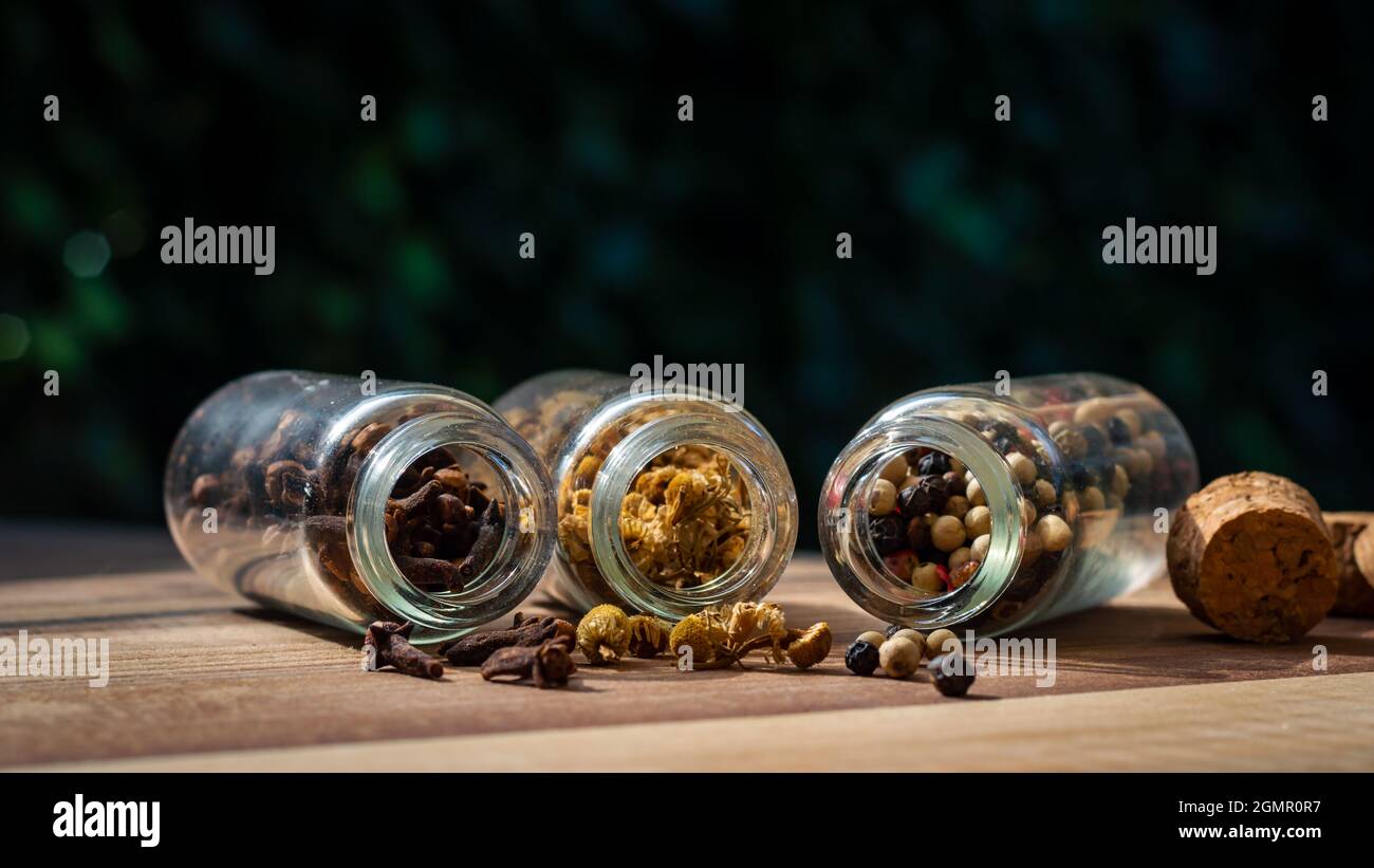 Ein Bild von drei Glasflaschen mit Pfefferkörnern, Kamille und Nelken, die auf einer Holzoberfläche auslaufen Stockfoto