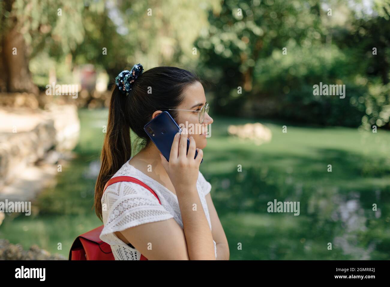 Mittellange Aufnahme einer jungen Frau mit einem weißen Kleid und einem roten Rucksack, die in einem Park telefoniert Stockfoto