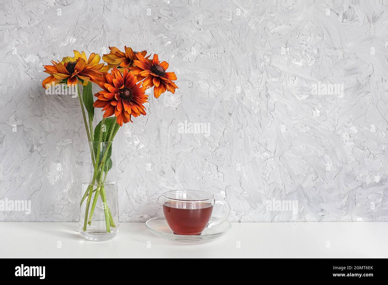 Teezeit. Schwarzer Tee in durchsichtiger Tasse mit Untertasse und Strauß orangefarbener Blumen Koneblüten in Vase auf Tisch gegen graue Steinwand mit Kopierraum M Stockfoto