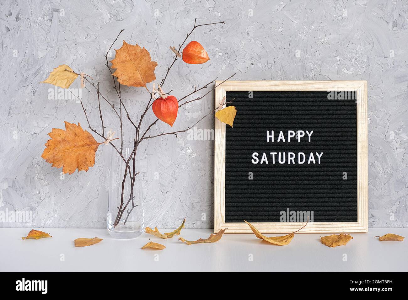 Happy Saturday Text auf schwarzem Briefbrett und Strauß von Zweigen mit gelben Blättern auf Wäscheklammern in Vase auf Tisch Vorlage für Postkarte, Gruß c Stockfoto