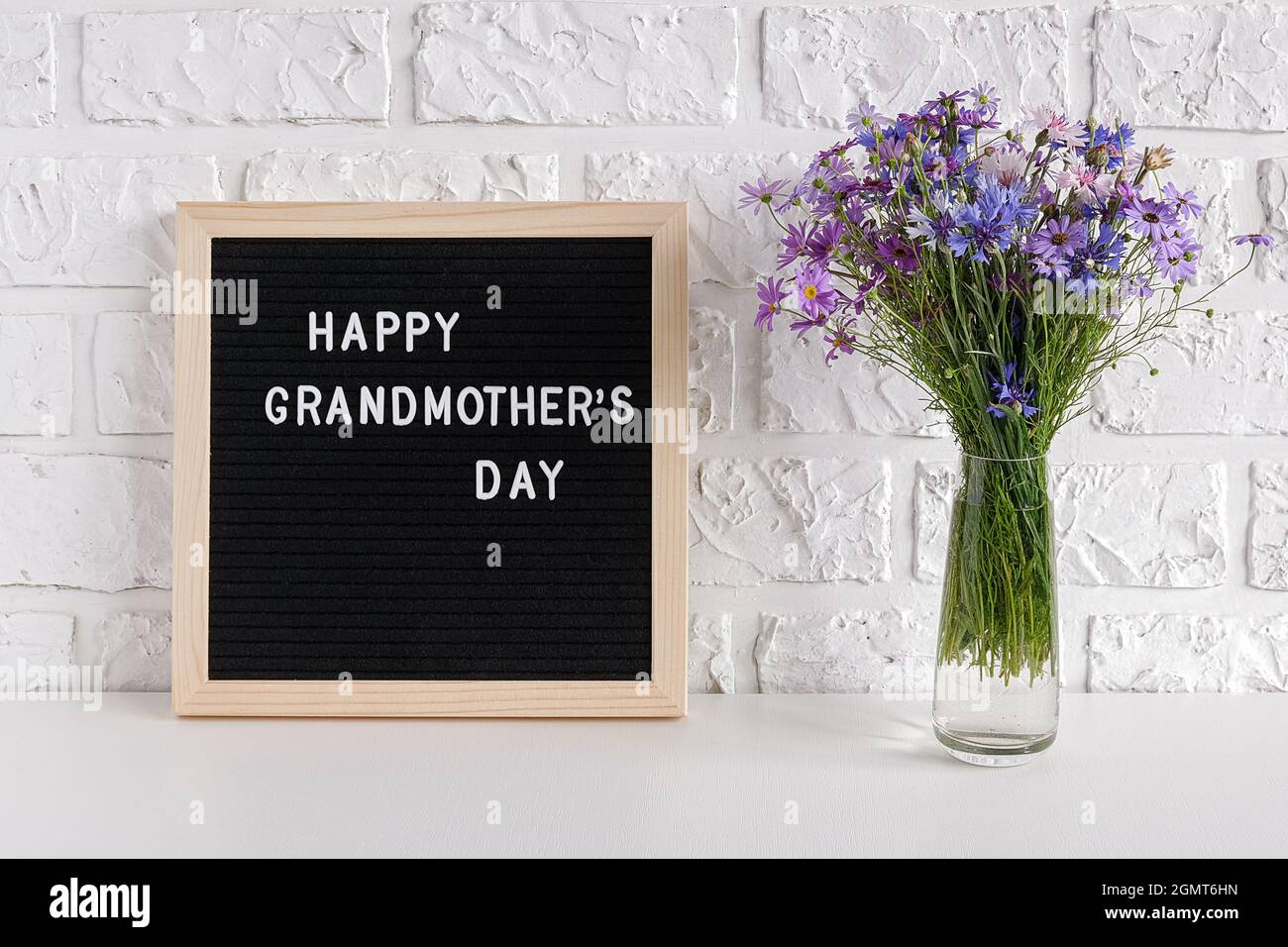 Fröhlicher Großmütter-Tag Text auf schwarzem Briefbrett und Blumenstrauß in blauer Vase auf dem Tisch gegen weiße Ziegelwand. Konzept Muttertag. Vorlage für Stockfoto